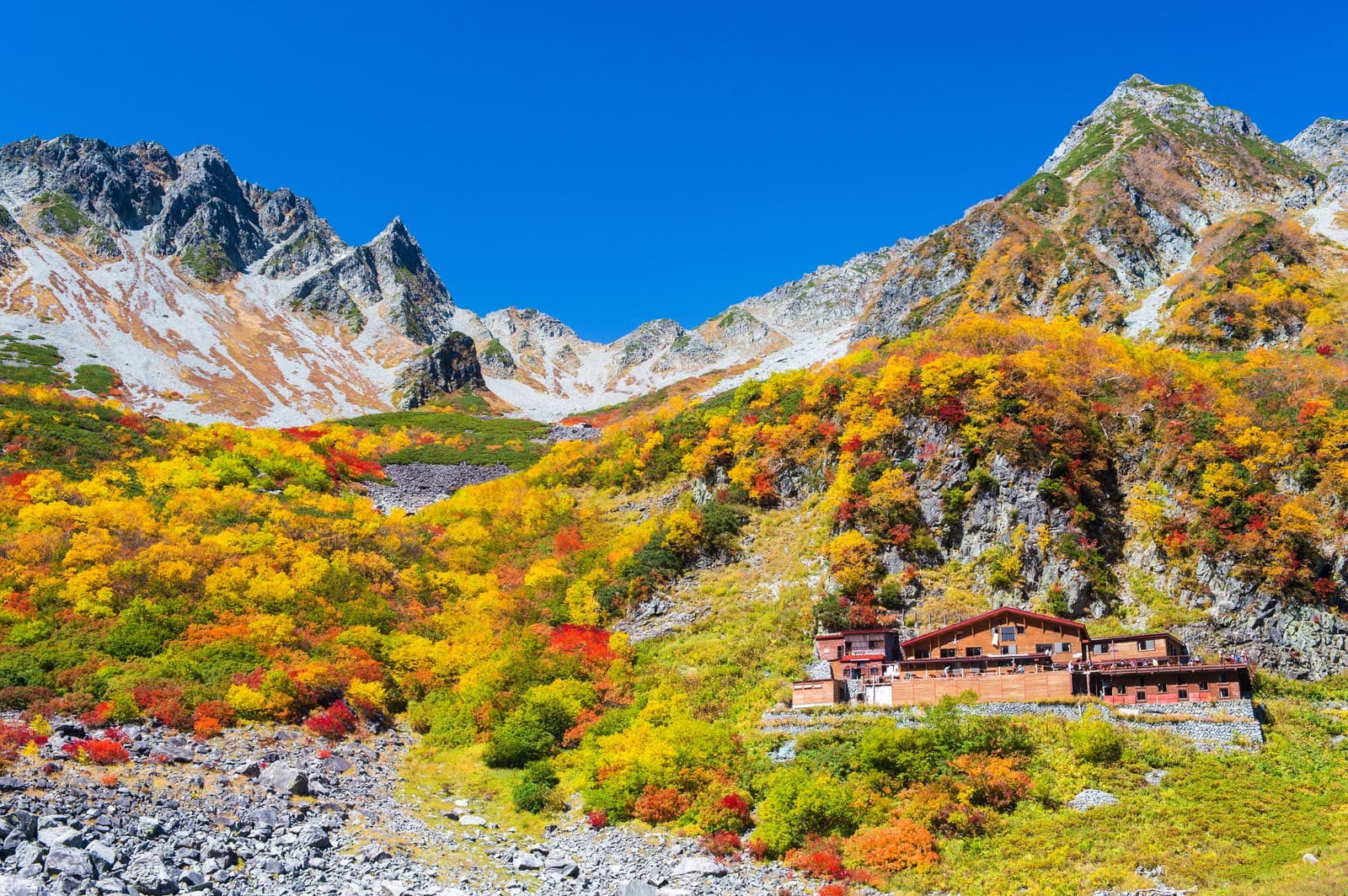 Red and yellow autumn foliage below the rocky peaks of Karasawa Cirque in the Northern Japan Alps, with a mountain hut on the slope